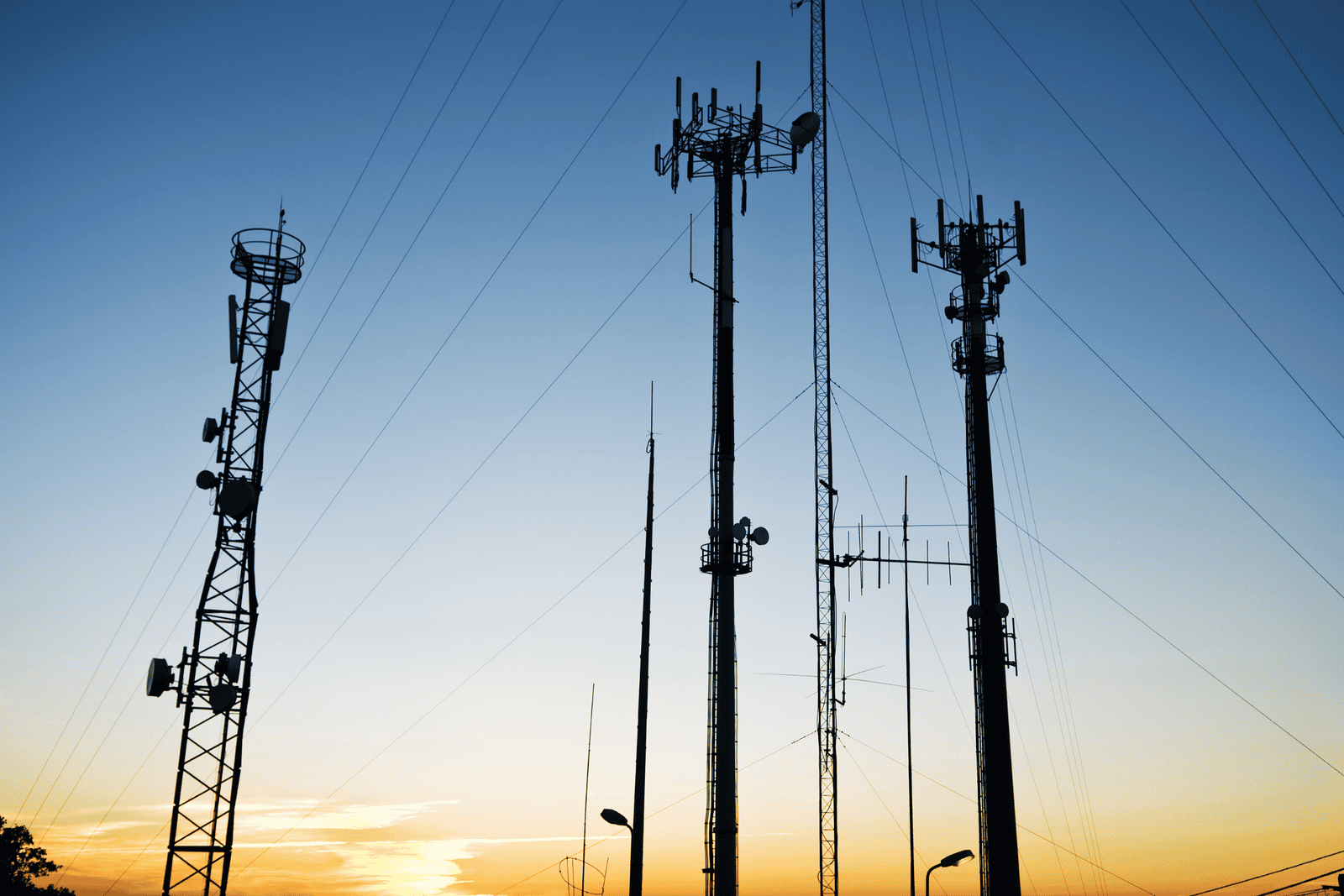 Silhouetted telecommunications towers with antennas and cables against a gradient sunset sky, transitioning from warm orange near the horizon to deep blue above.